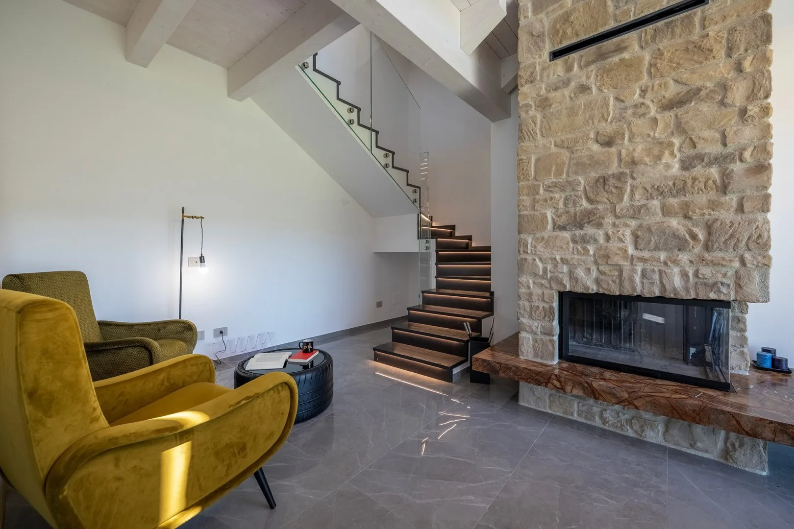 Una sala de estar moderna cuenta con una chimenea de piedra, una escalera de madera oscura y sillones amarillo mostaza.