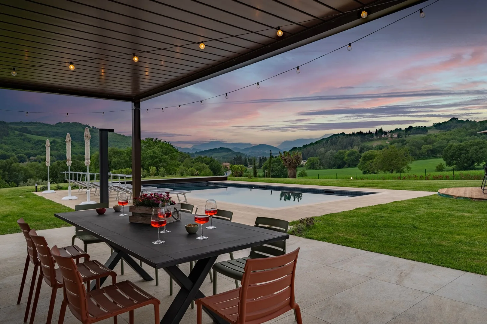 Una terraza cubierta con comedor da a una piscina, colinas verdes y un cielo colorido al atardecer.