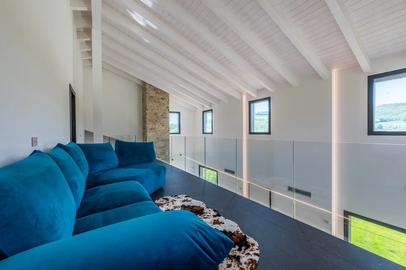 Sala tipo loft con un sofá seccional azul, suelo oscuro, barandilla de vidrio y techo alto con vigas blancas.