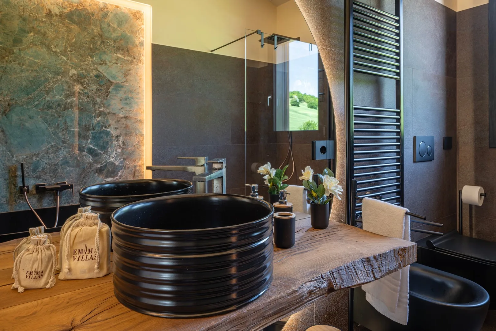 Un baño moderno cuenta con dos lavabos negros sobre un tocador de madera, una ducha de vidrio y una bañera independiente.