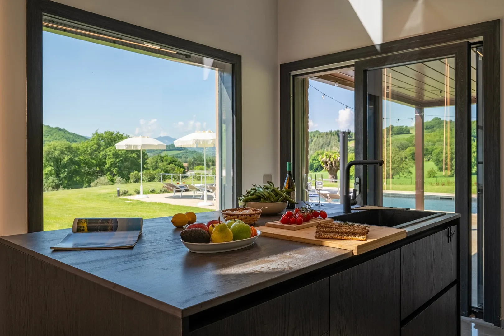 Una isla de cocina moderna con fruta y tablas de cortar da a un jardín verde a través de grandes ventanales.