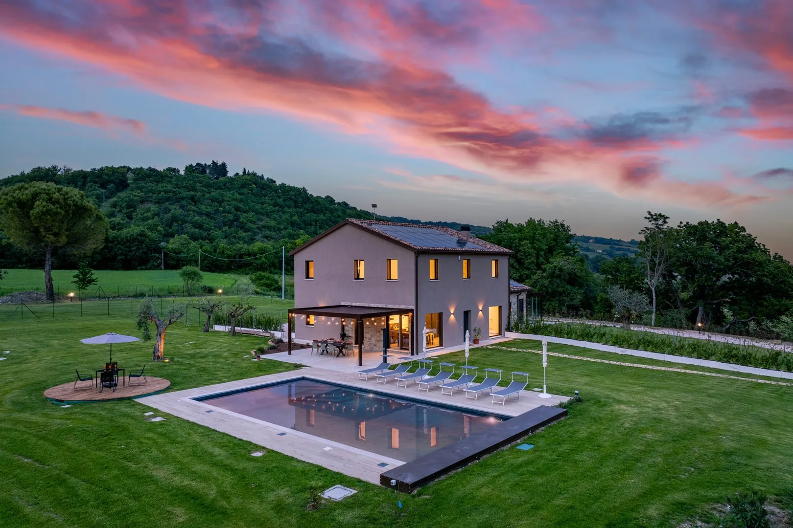 Una casa moderna de dos plantas se alza junto a una piscina rectangular en un amplio césped, bajo un colorido cielo vespertino.