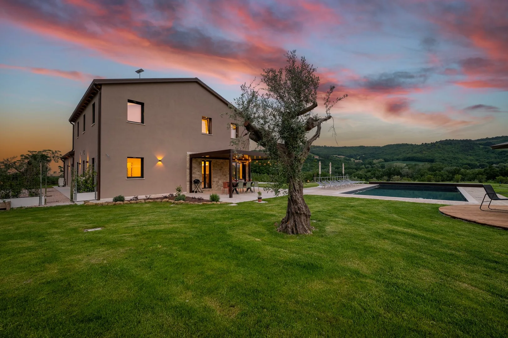 Una casa moderna de dos plantas con césped, piscina y olivo bajo un cielo de atardecer colorido.