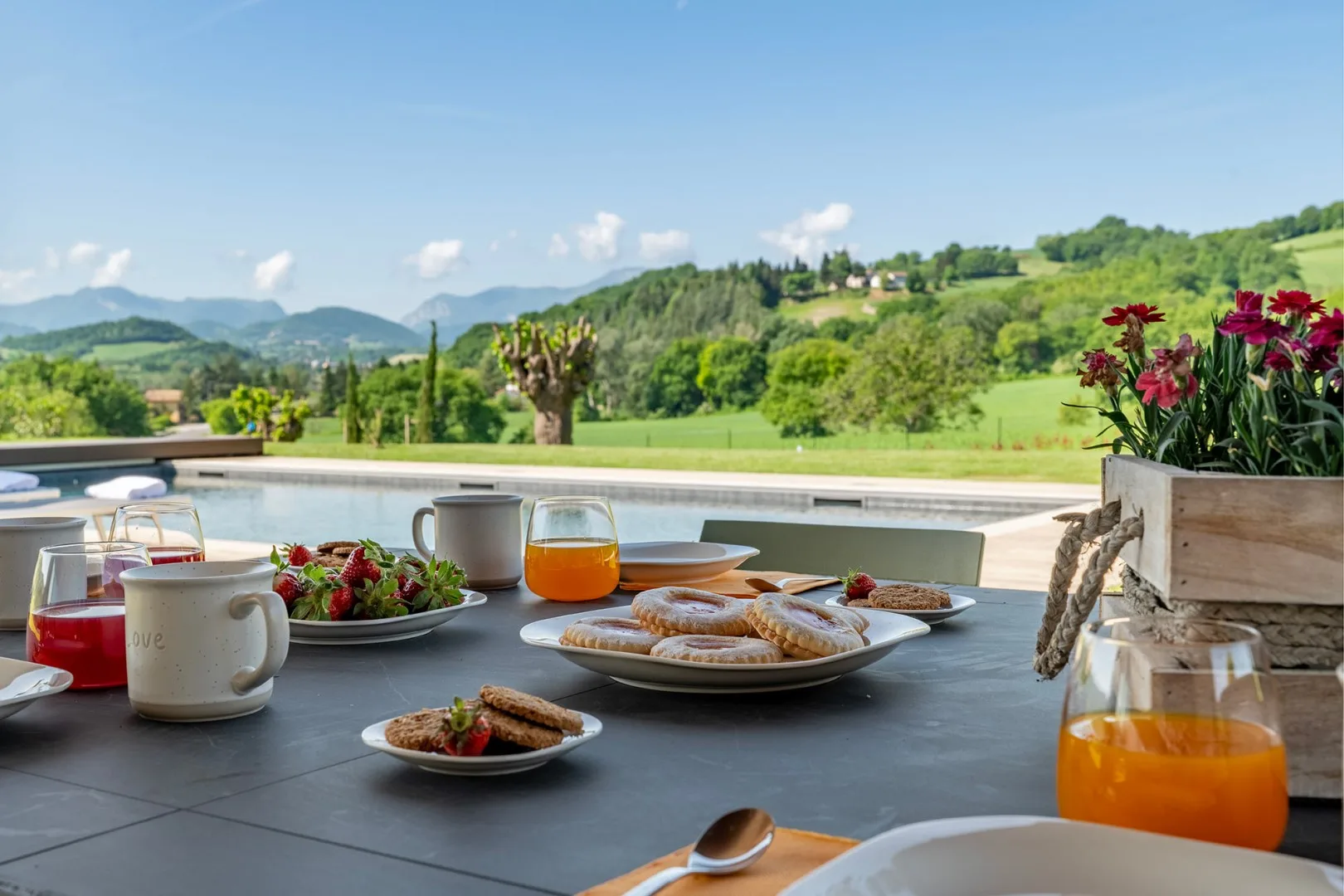 Una mesa de desayuno con zumo, café, bollería y fruta da a colinas verdes y montañas lejanas.