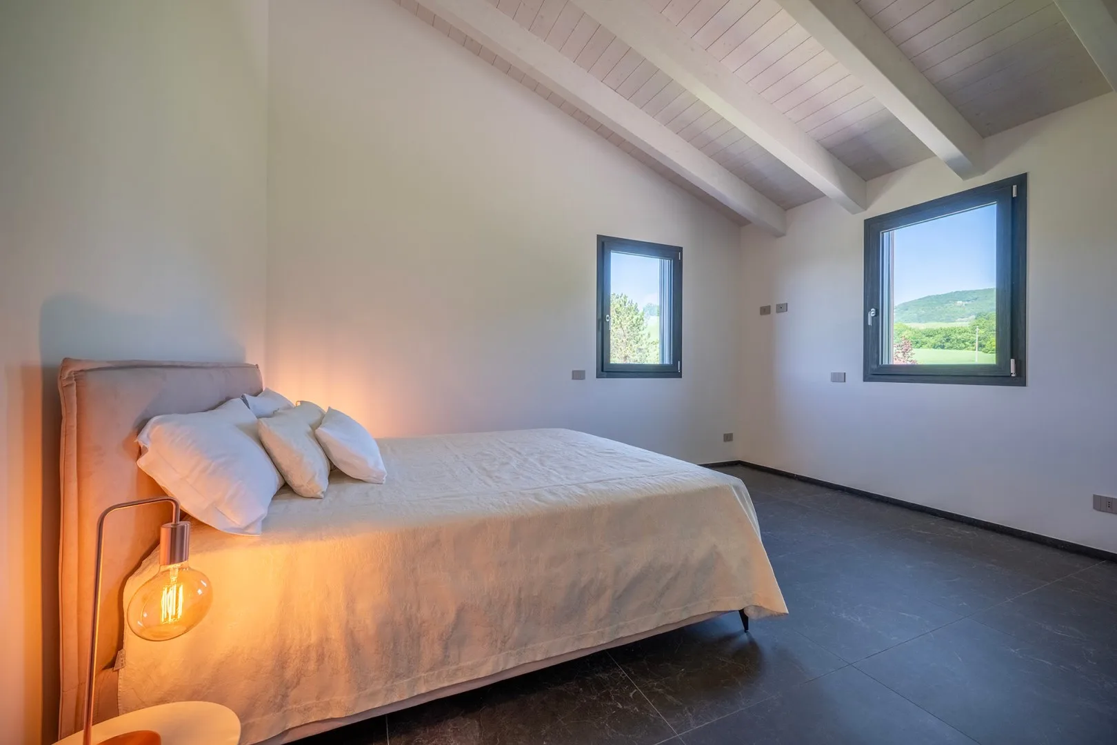 Un dormitorio con una cama bien tendida, techo inclinado con vigas y dos ventanas con vistas a colinas verdes.