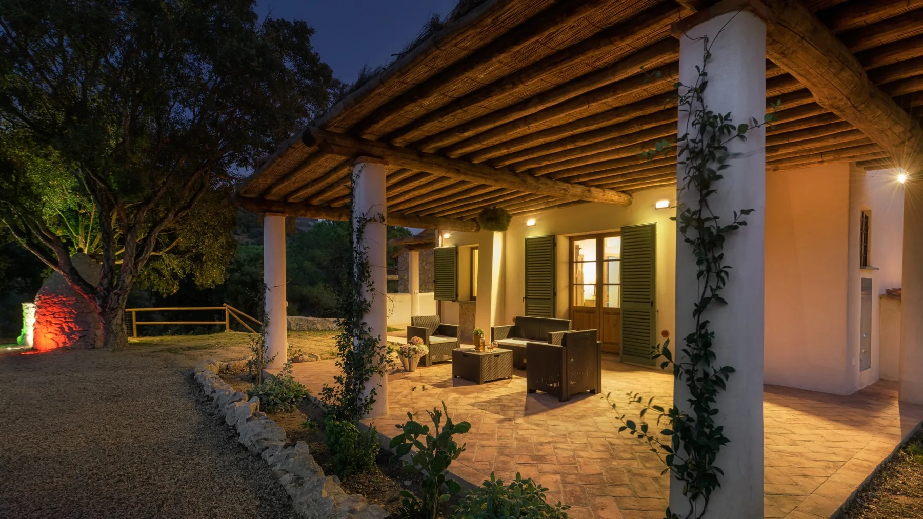Un patio exterior cubierto con asientos, ventanas iluminadas y un camino de grava.