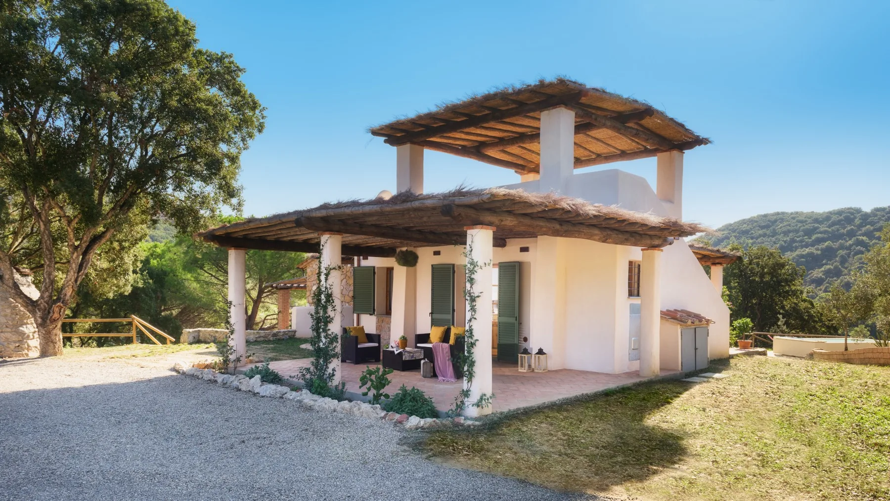 Una casa blanca con un patio sombreado está junto a un camino de grava.