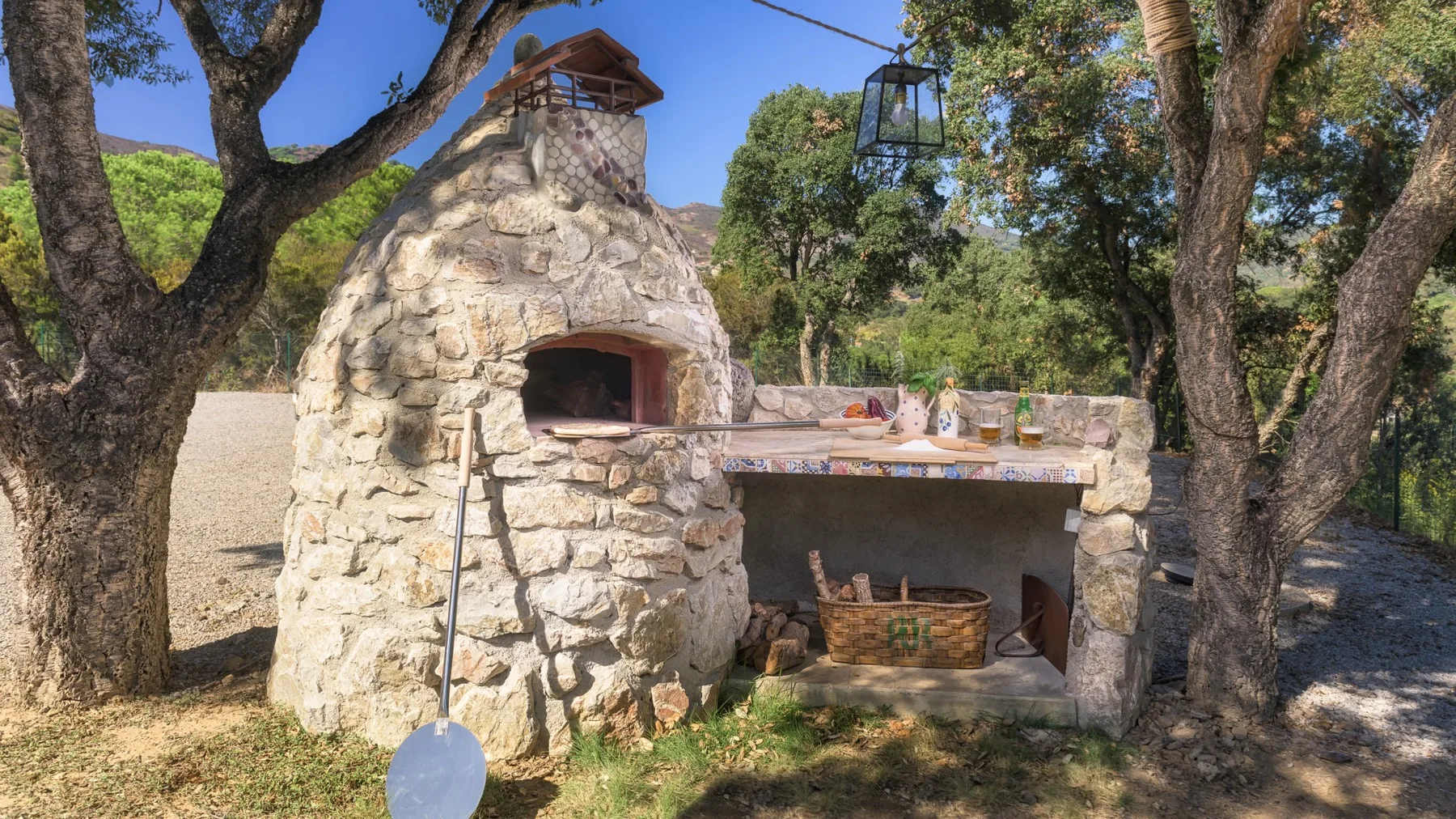 Un horno de piedra al aire libre y una zona de cocina contigua entre árboles.