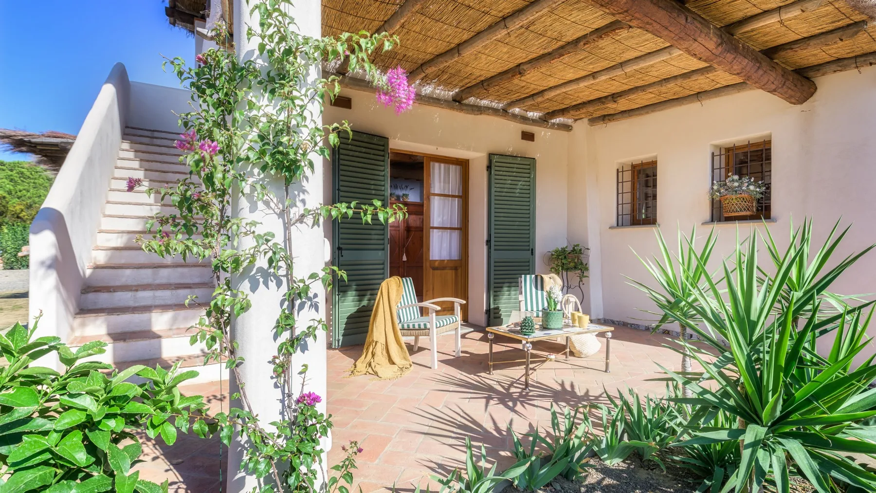 Un patio sombreado con asientos, contraventanas verdes y plantas trepadoras con flores.