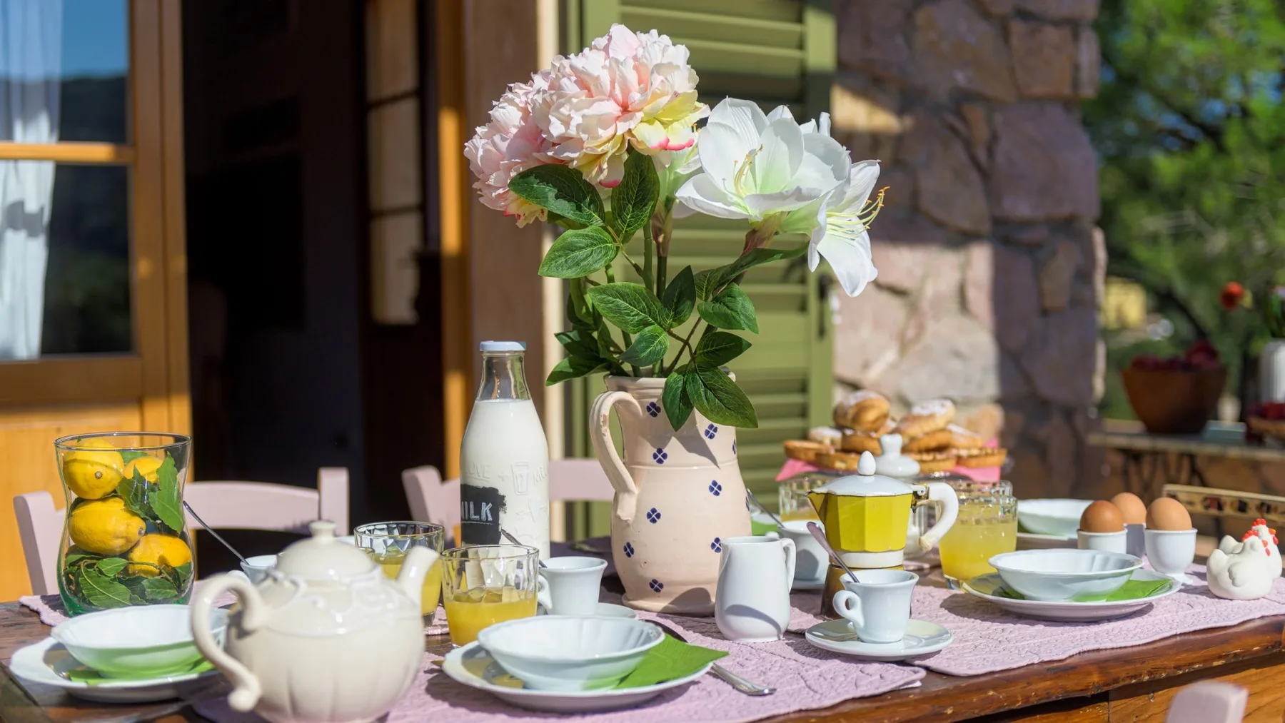 Una mesa al aire libre está preparada con platos, bebidas y un jarrón de flores.