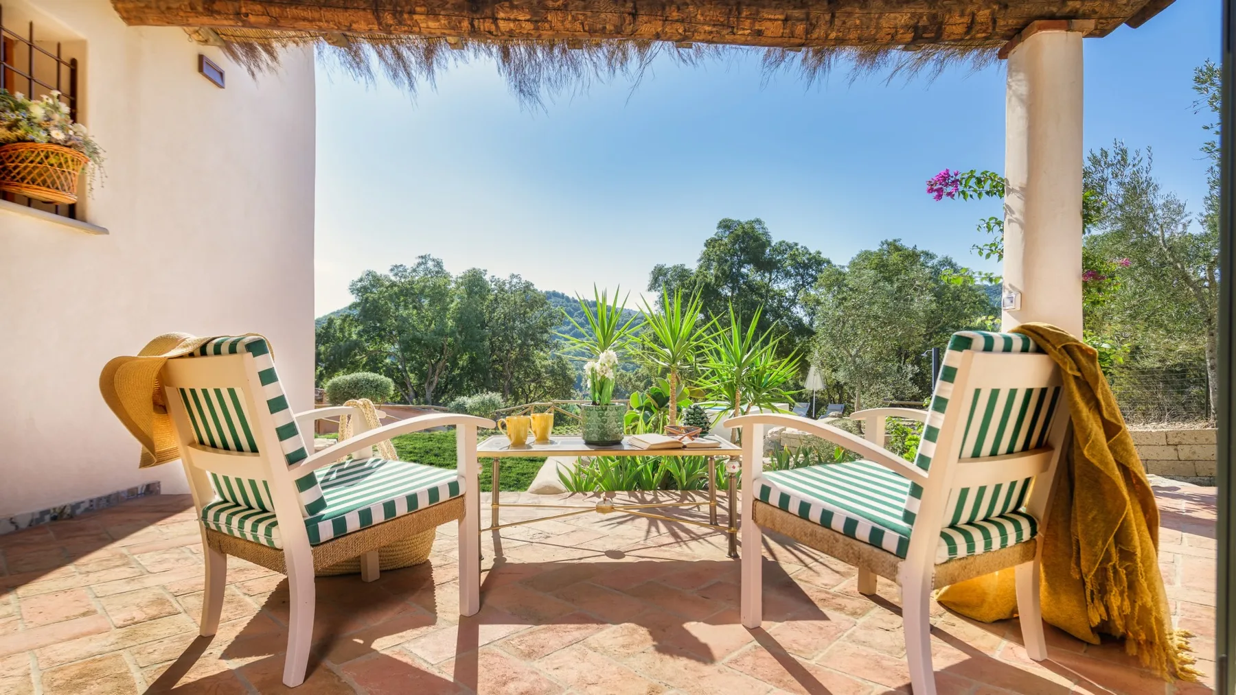 Una zona de estar exterior a la sombra da a un jardín con árboles y plantas en macetas.
