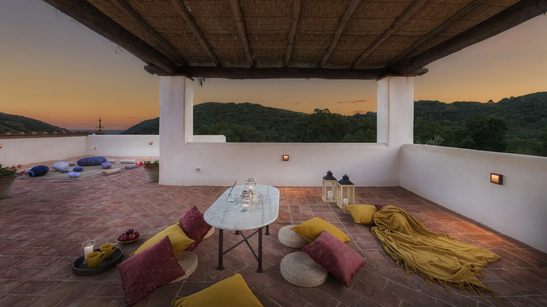 Terraza cubierta con cojines en el suelo, una mesa baja y vistas a colinas verdes al atardecer.
