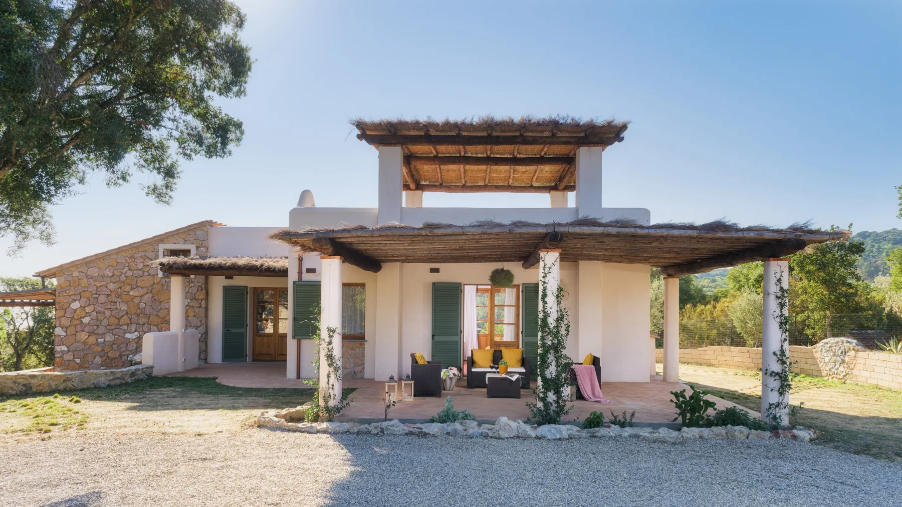 Una casa baja con un muro lateral de piedra y un patio cubierto se alza en un patio de grava.