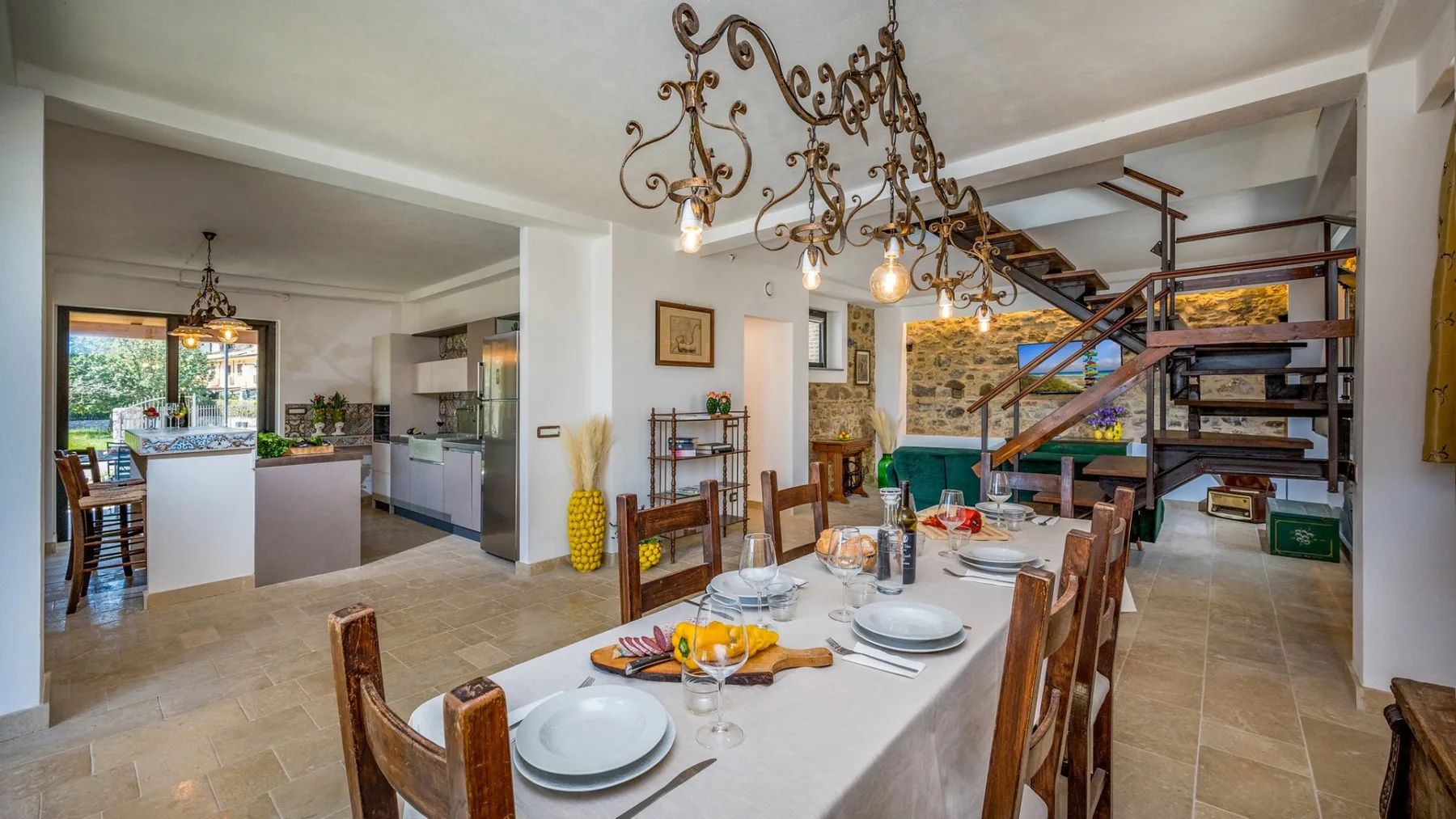 Una zona abierta de comedor y cocina tiene una mesa larga puesta bajo lámparas colgantes ornamentadas.