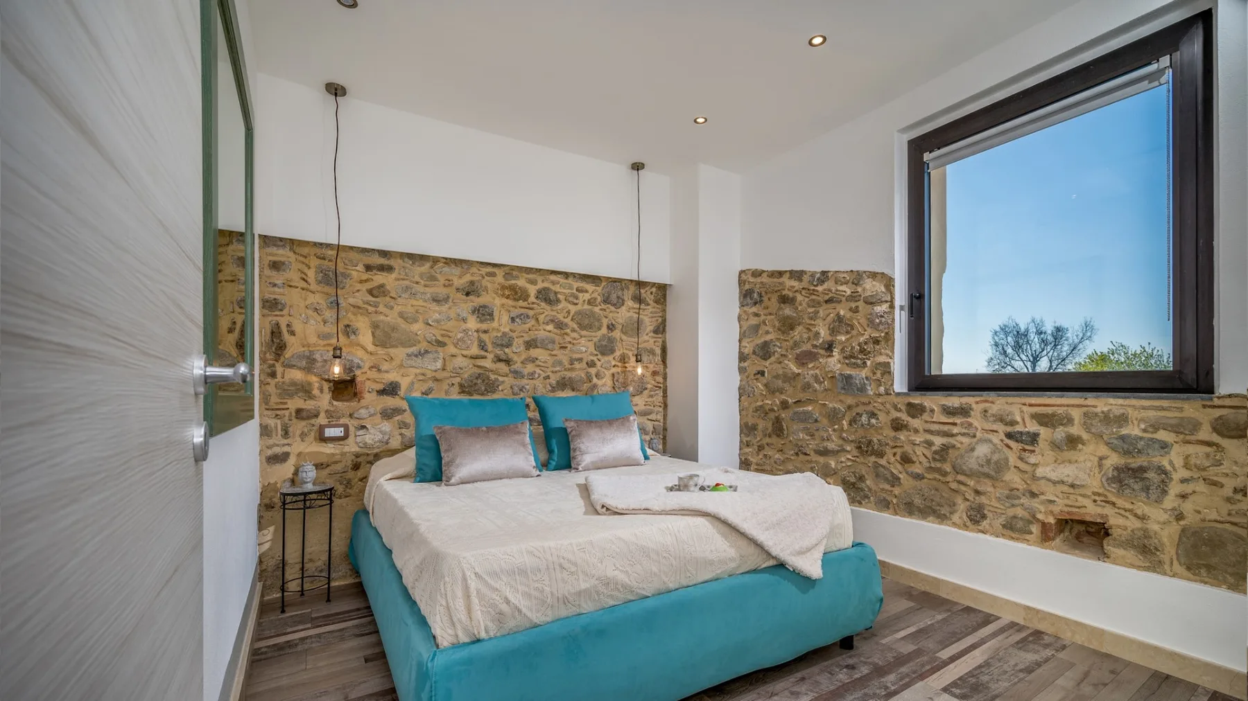 Dormitorio con una cama turquesa, ropa de cama clara y paredes de piedra bajo una ventana grande.