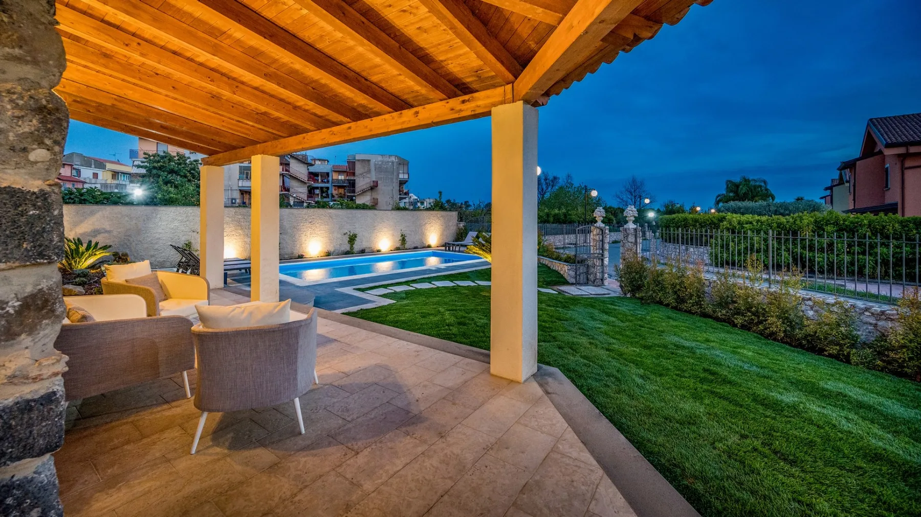Un patio cubierto con asientos exteriores da a una piscina iluminada y un césped al anochecer.