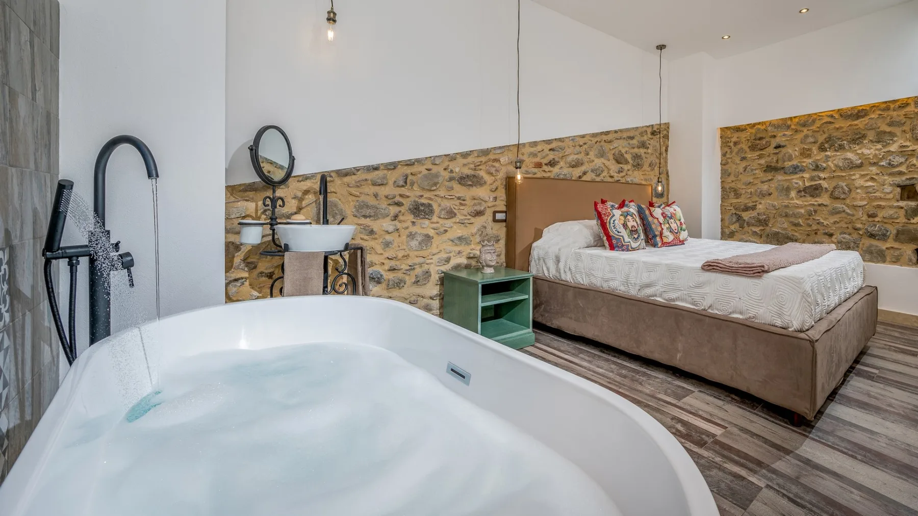 Un dormitorio con una bañera grande en primer plano y una cama junto a una pared de piedra.