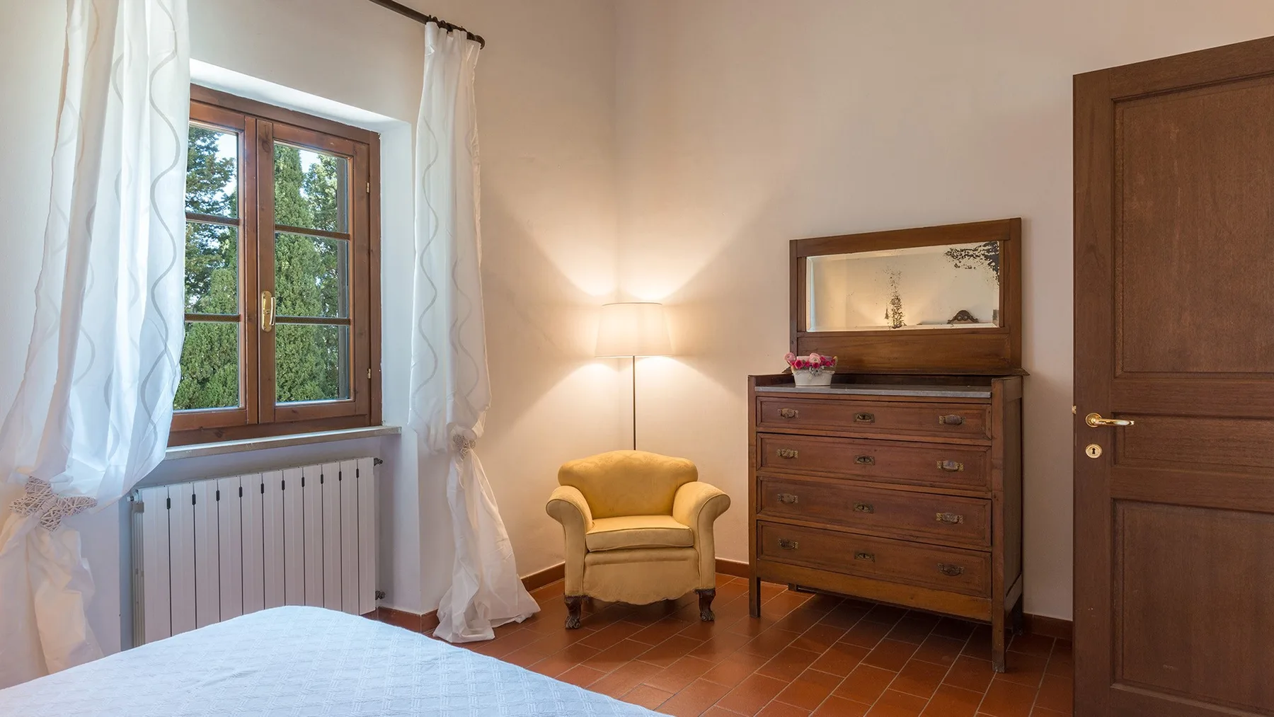 Un dormitorio con una ventana, un sillón y una cómoda de madera.
