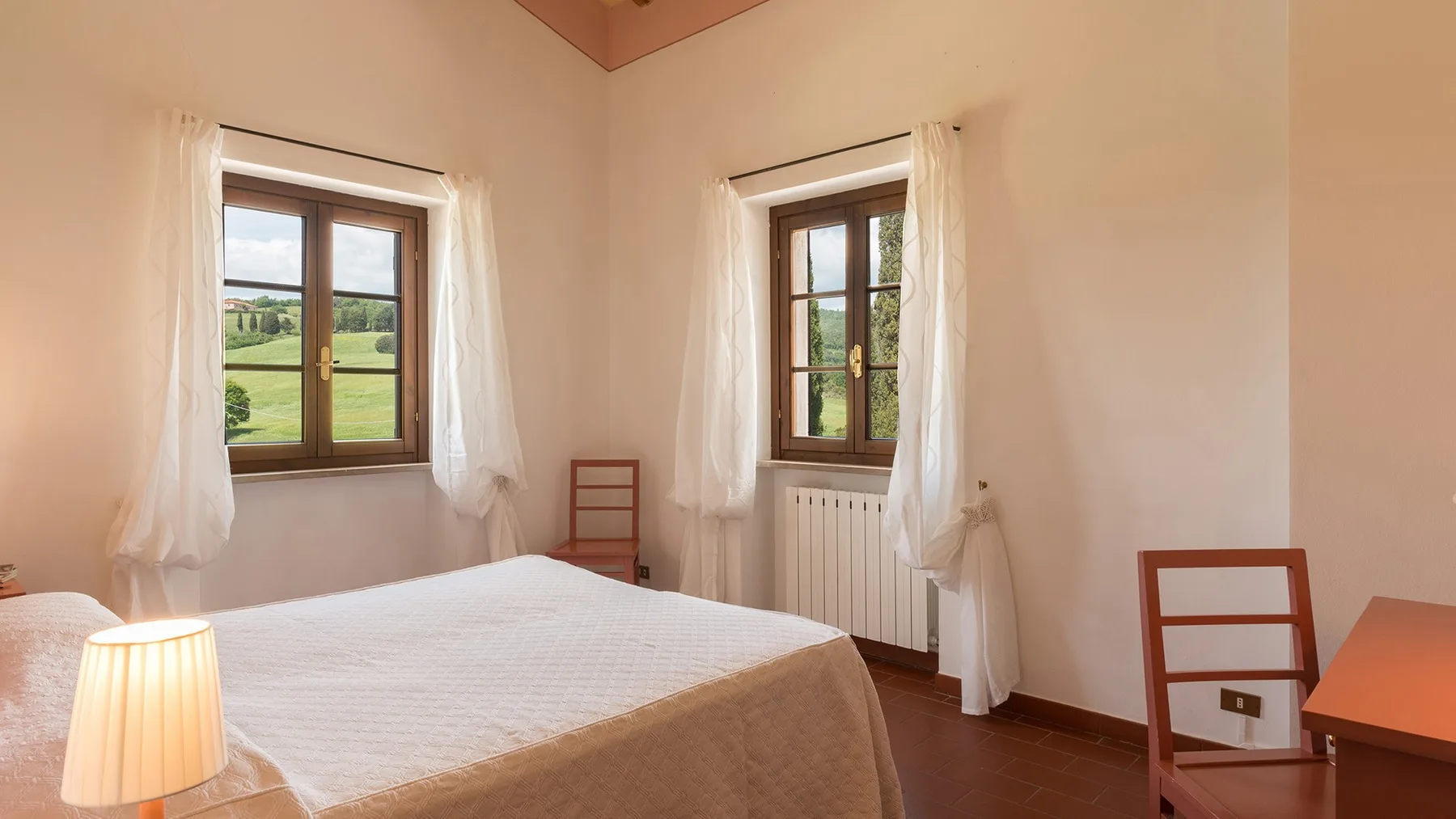 Un dormitorio con una cama bien tendida y dos ventanas con vistas a campos verdes.