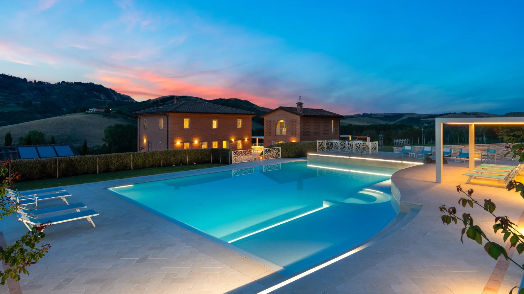 An outdoor swimming pool glows beside a paved terrace at dusk.