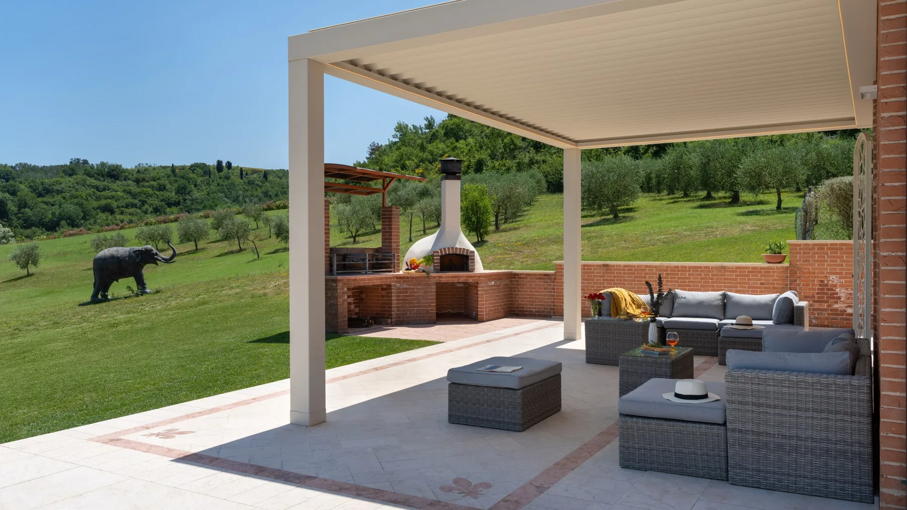A covered outdoor patio with seating overlooks a grassy, tree-lined landscape.