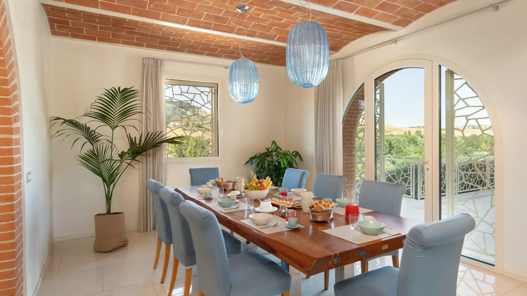 A dining area with a long table set for a meal beside large arched glass doors.