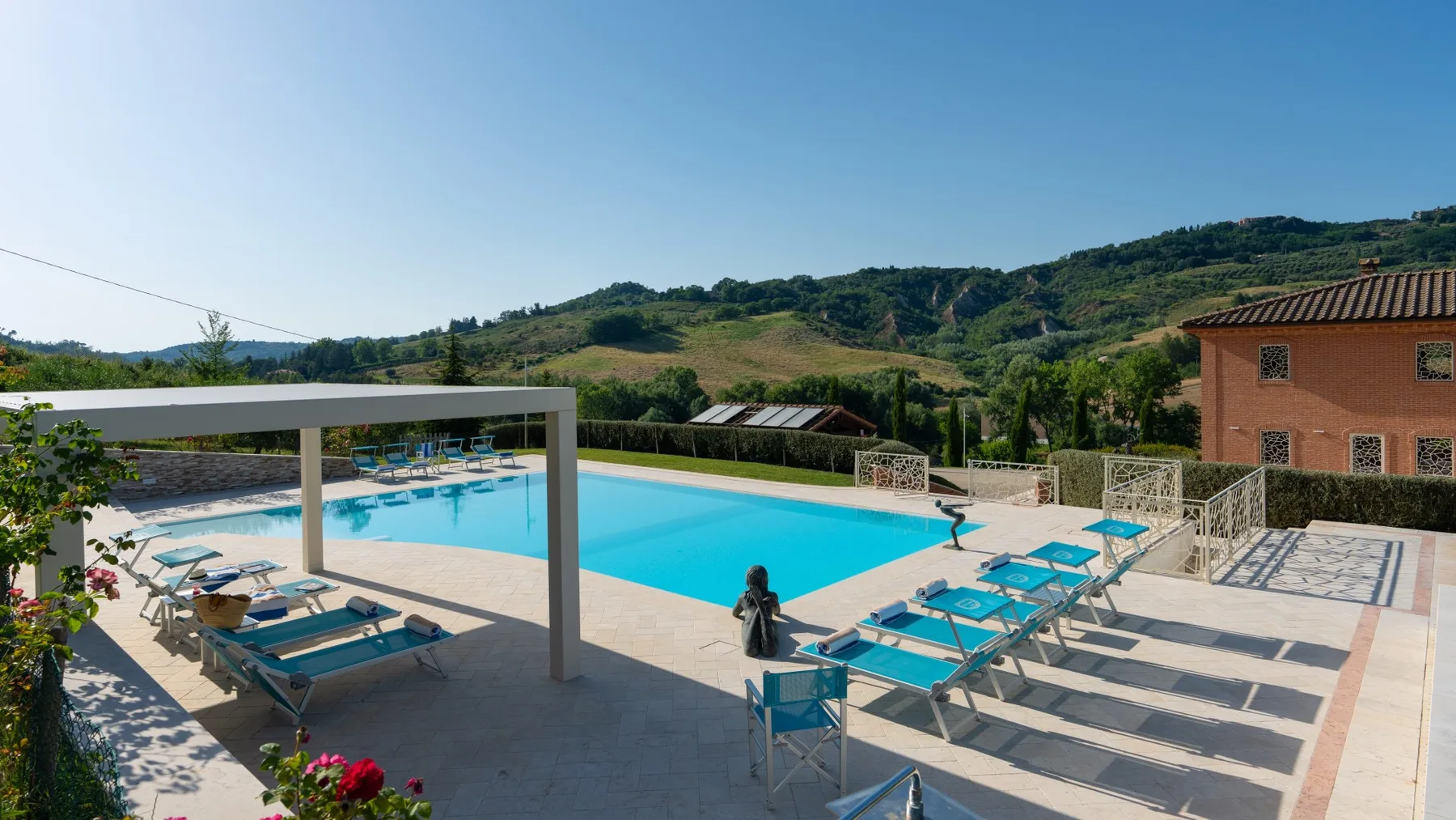 An outdoor swimming pool is surrounded by sun loungers and green hills.
