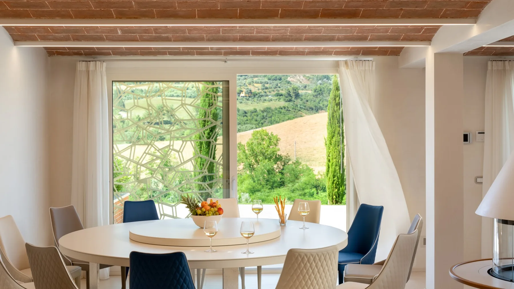 A dining area with a round table and large windows overlooking green countryside.
