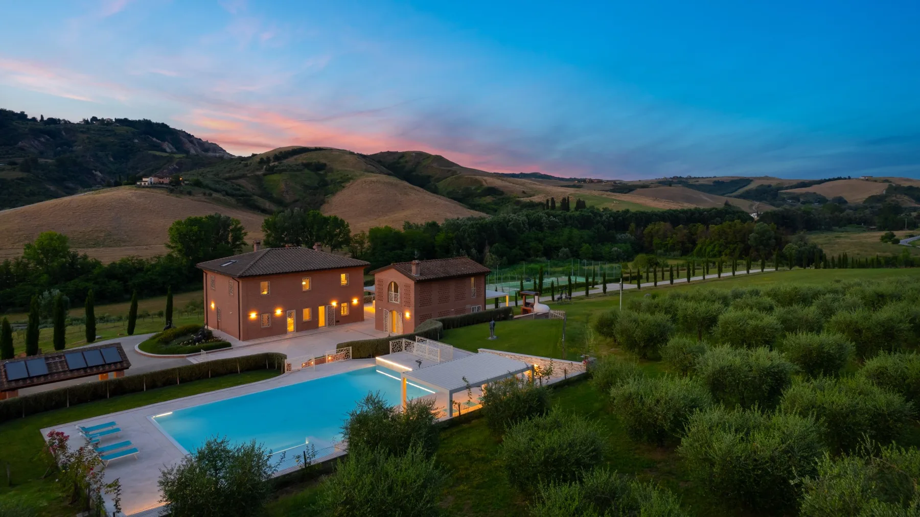 A countryside property with a lit building and swimming pool beneath rolling hills at dusk.