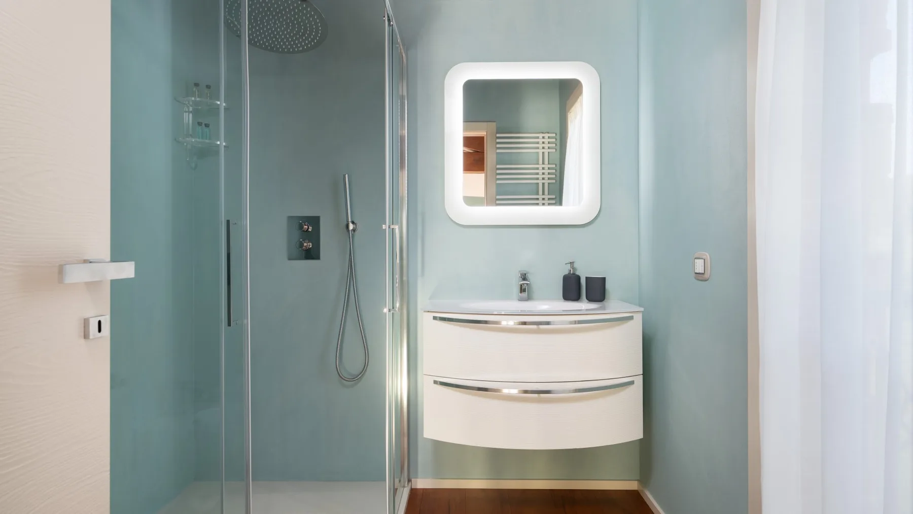 A bathroom with a glass shower and a curved vanity beneath an illuminated mirror.