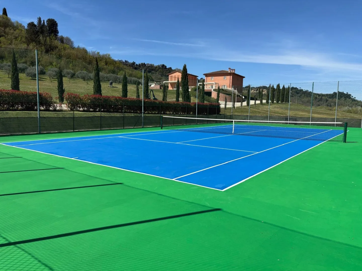 An outdoor tennis court with a blue playing surface and green surround.