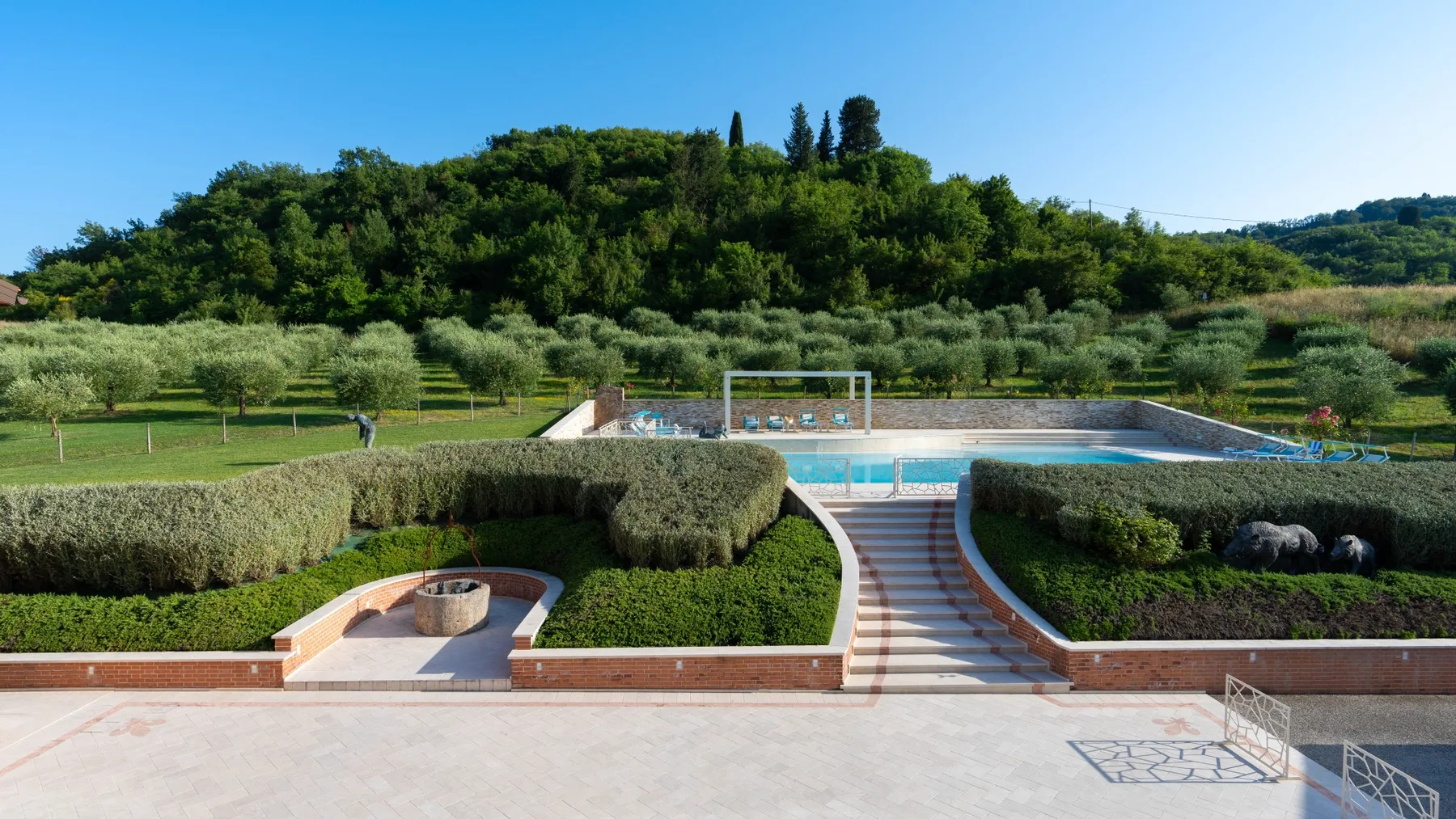 A landscaped outdoor swimming pool sits among trimmed hedges, with wooded hills beyond.