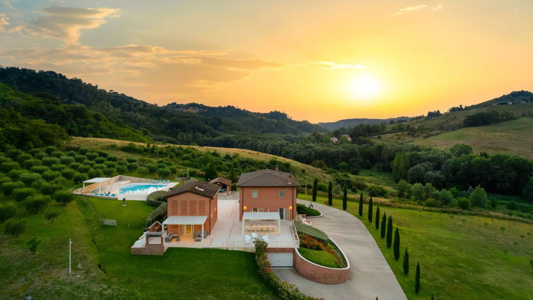 A countryside home sits among green hills beneath a glowing sunset.