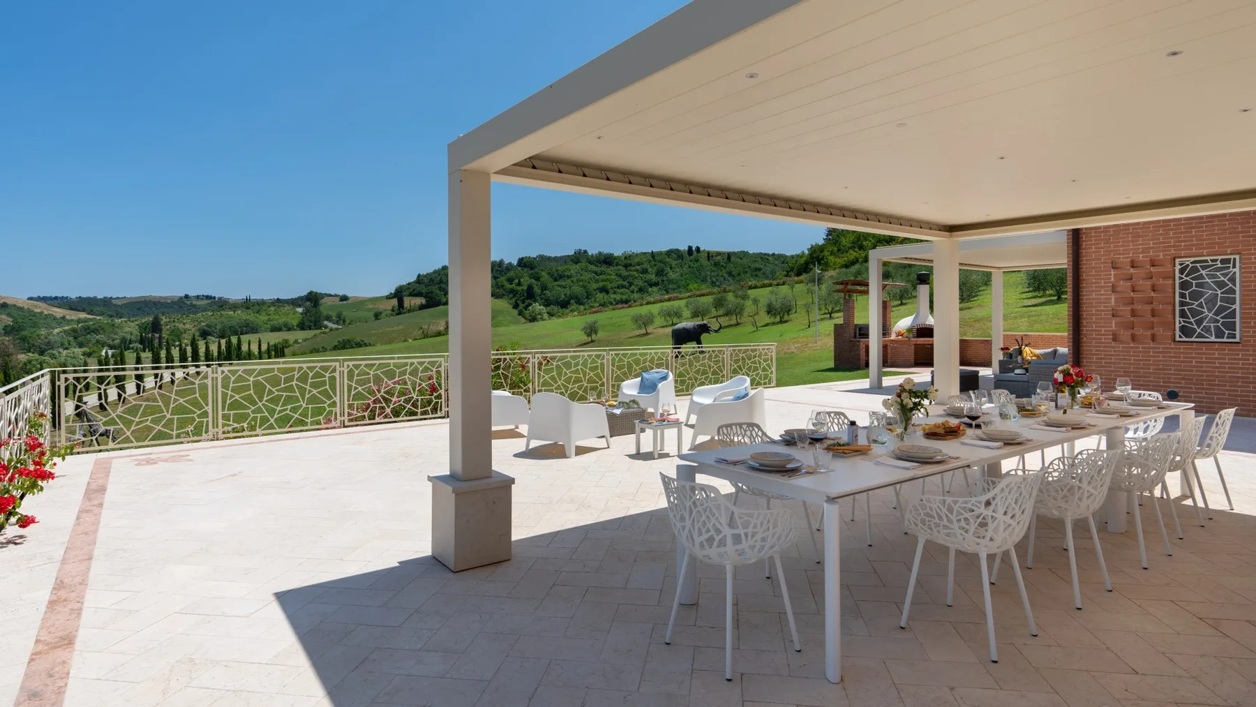 An outdoor dining terrace with a long table beneath a covered patio, overlooking green hills.