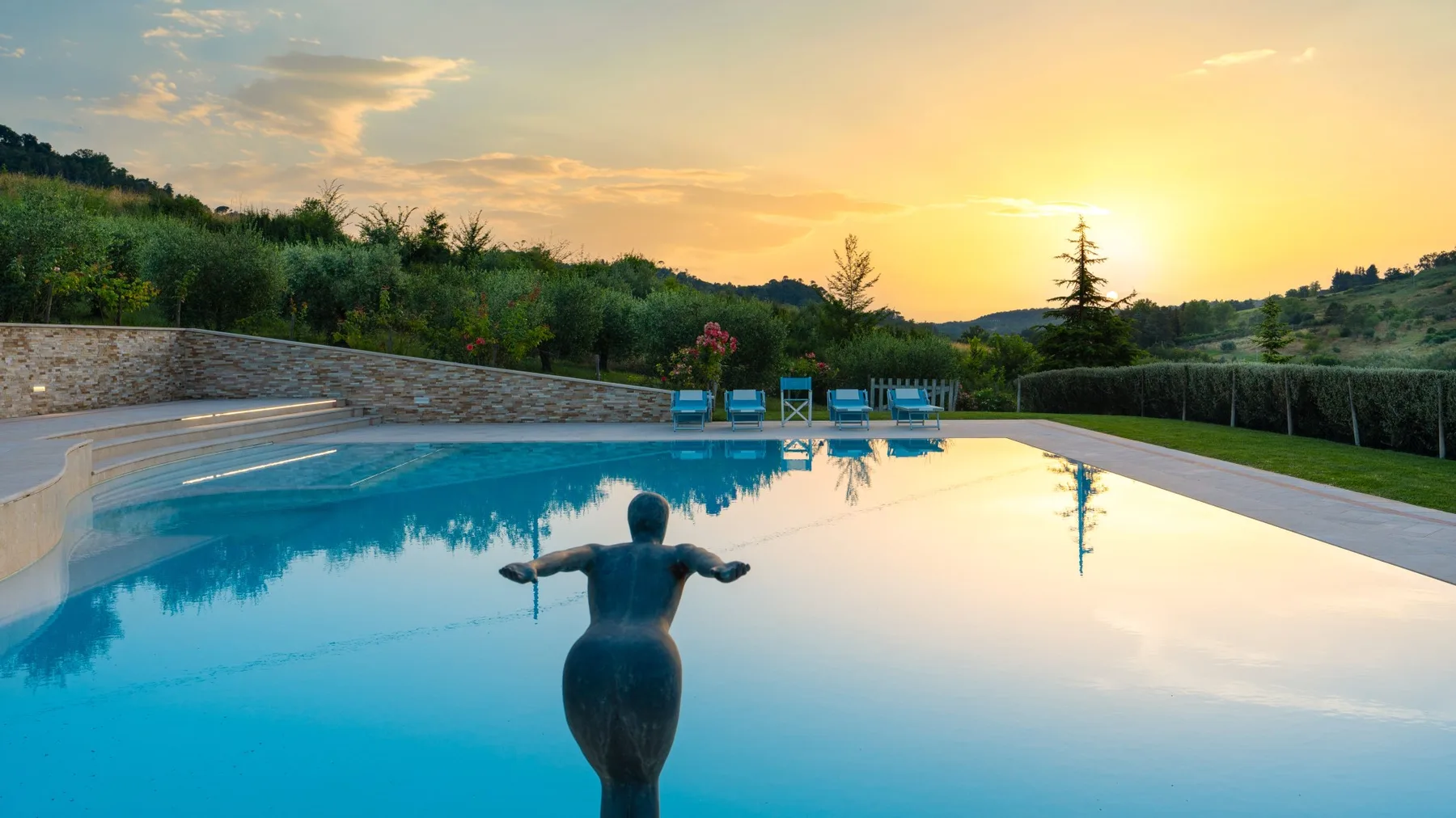 An outdoor swimming pool reflects the sunset, with a person standing in the water.