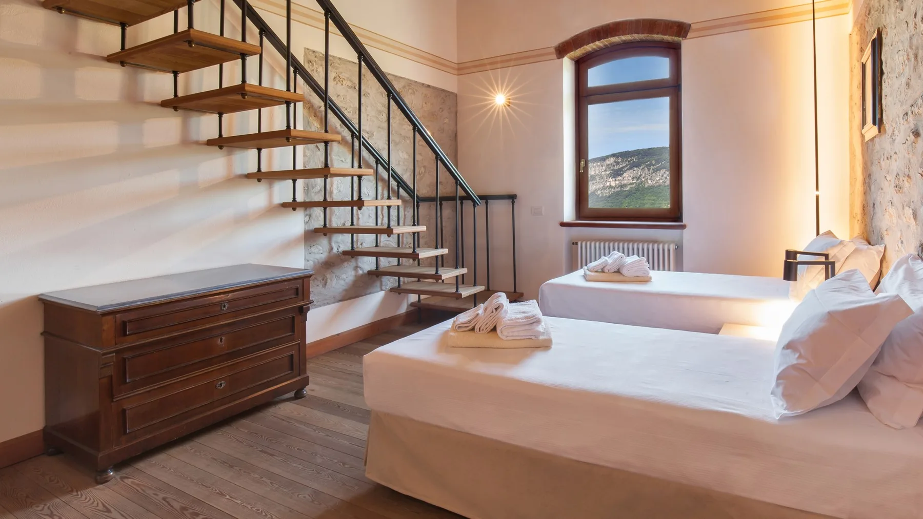 A bedroom with two beds, a wooden dresser, and a staircase beside a window.