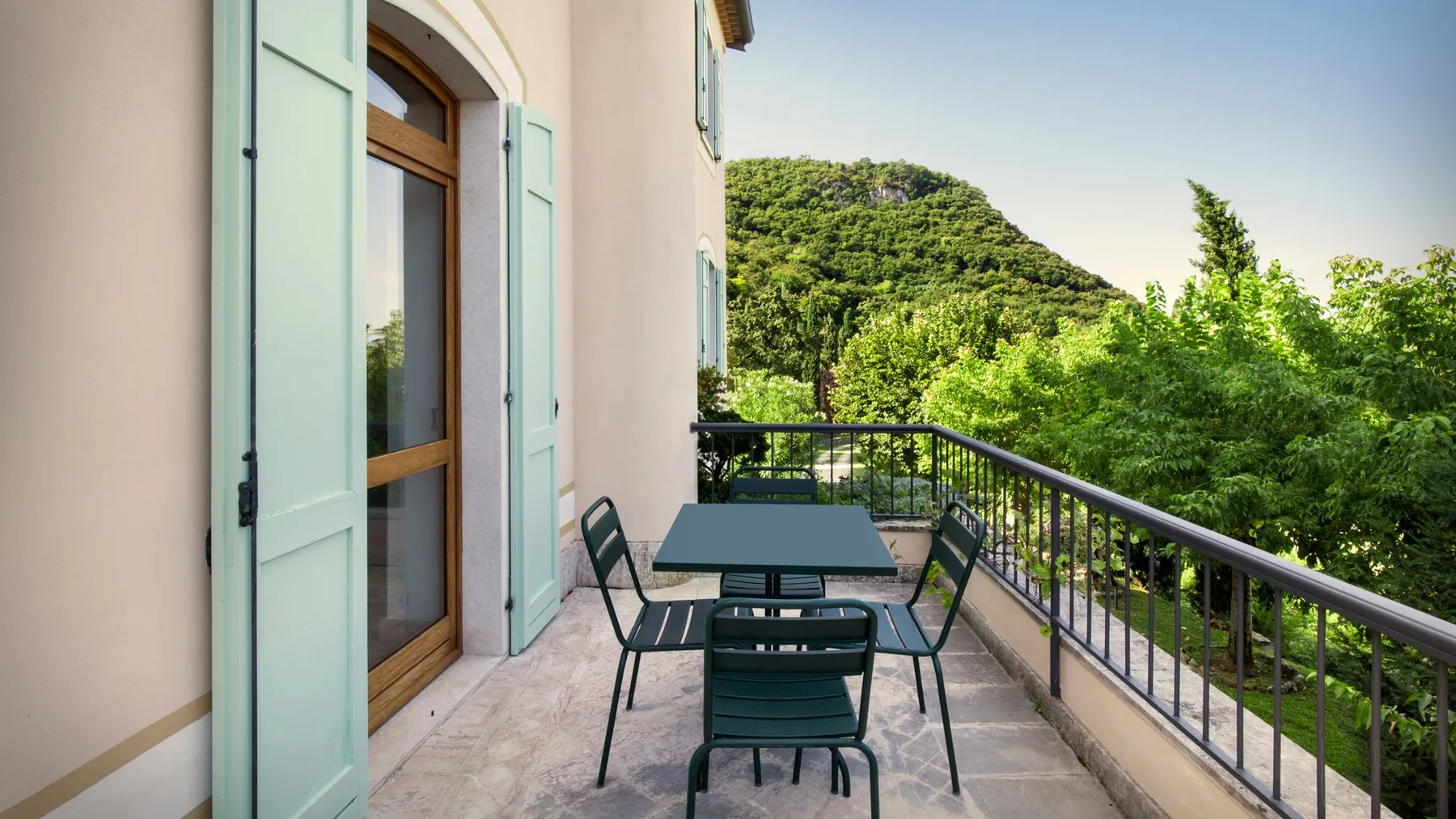 A tiled balcony has a table and seating beside a black railing overlooking green hills.