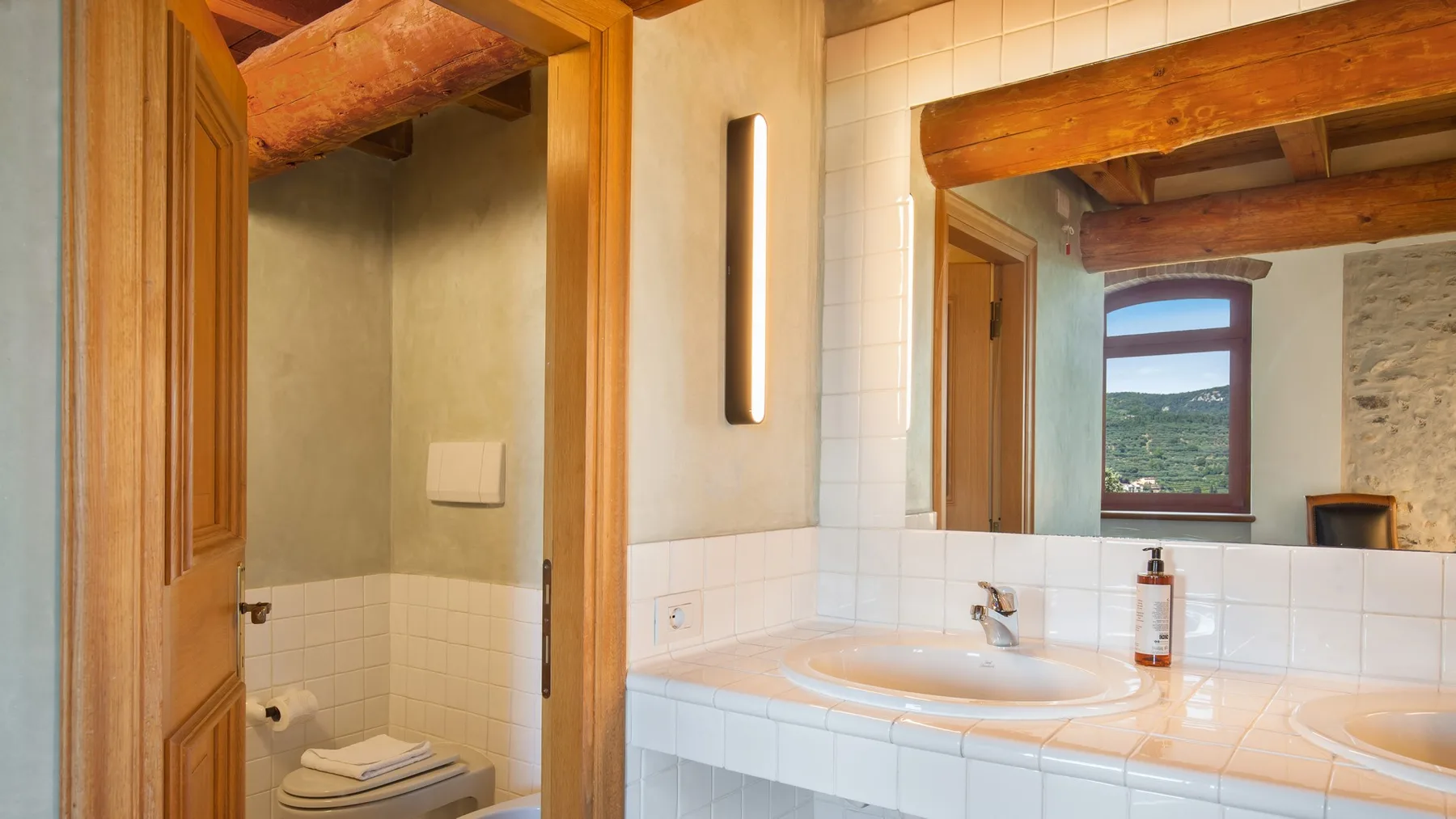 A tiled bathroom with a sink, mirror, and an open doorway to a toilet area.