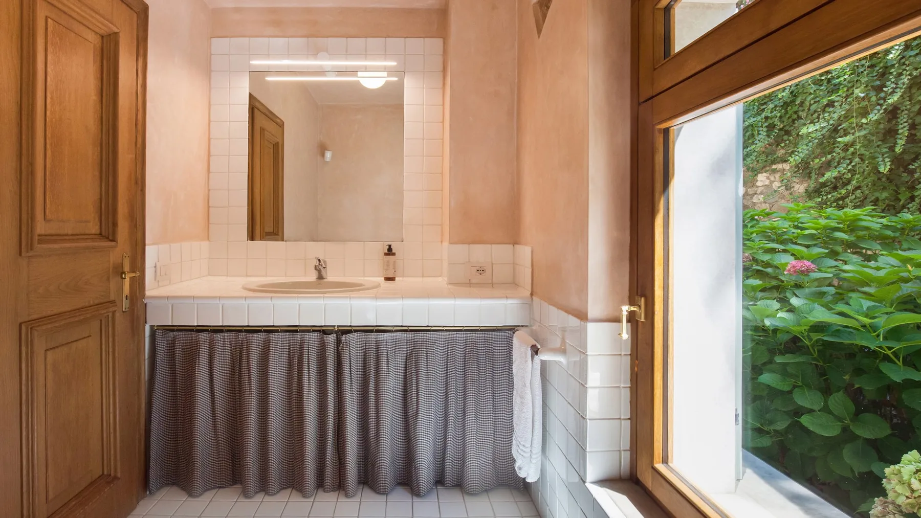 A bathroom vanity with a curtained cabinet and a window overlooking leafy greenery.