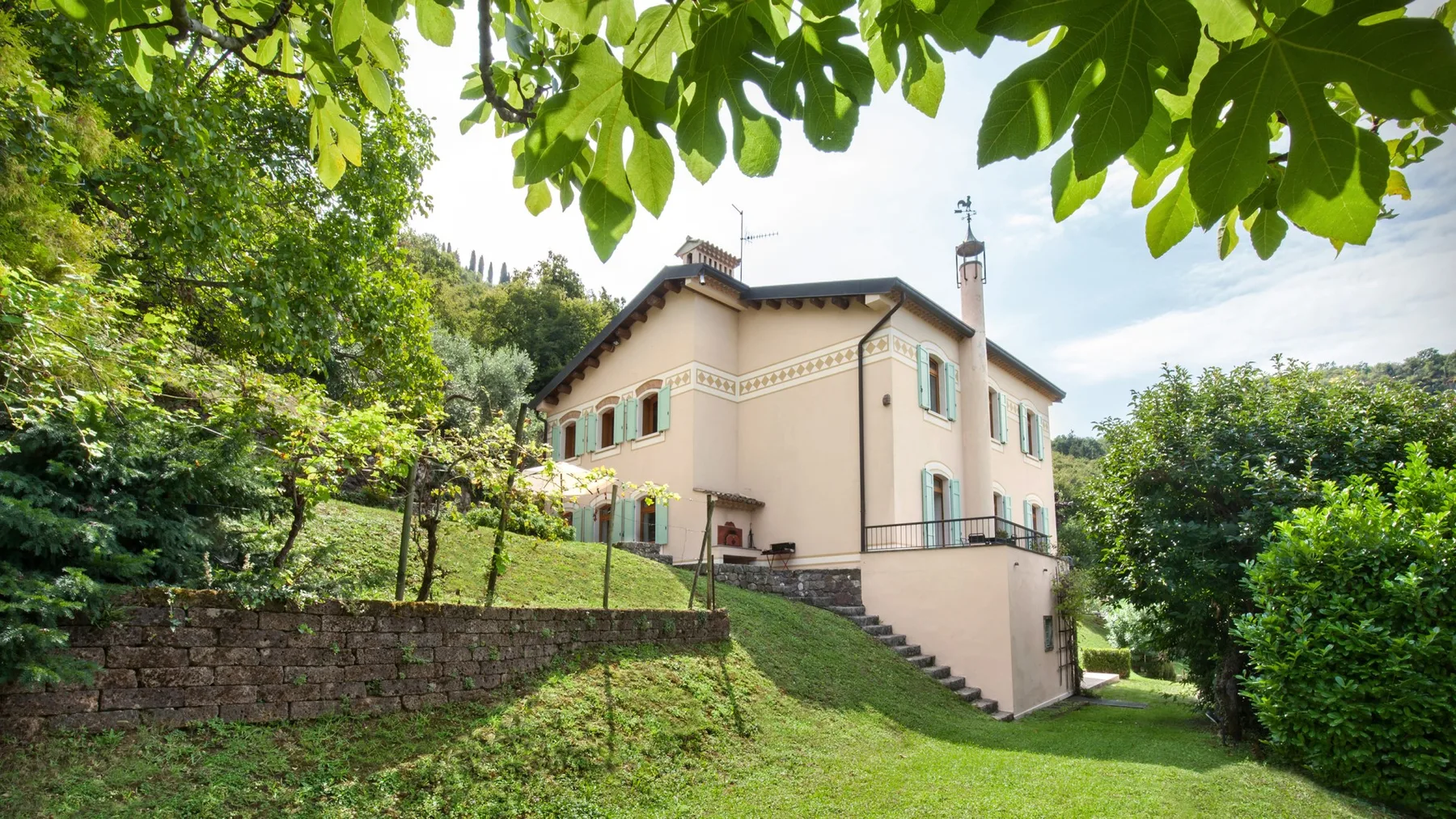 A pale two-story house stands on a grassy slope among trees.