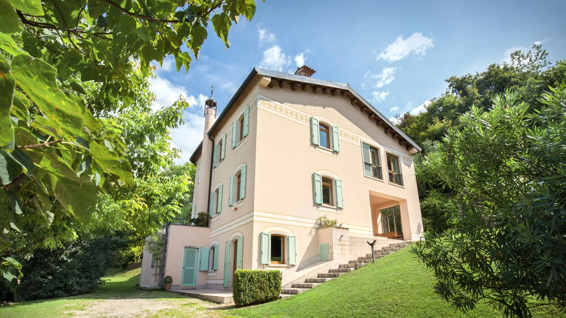 A pale house stands on a grassy slope, surrounded by leafy trees.