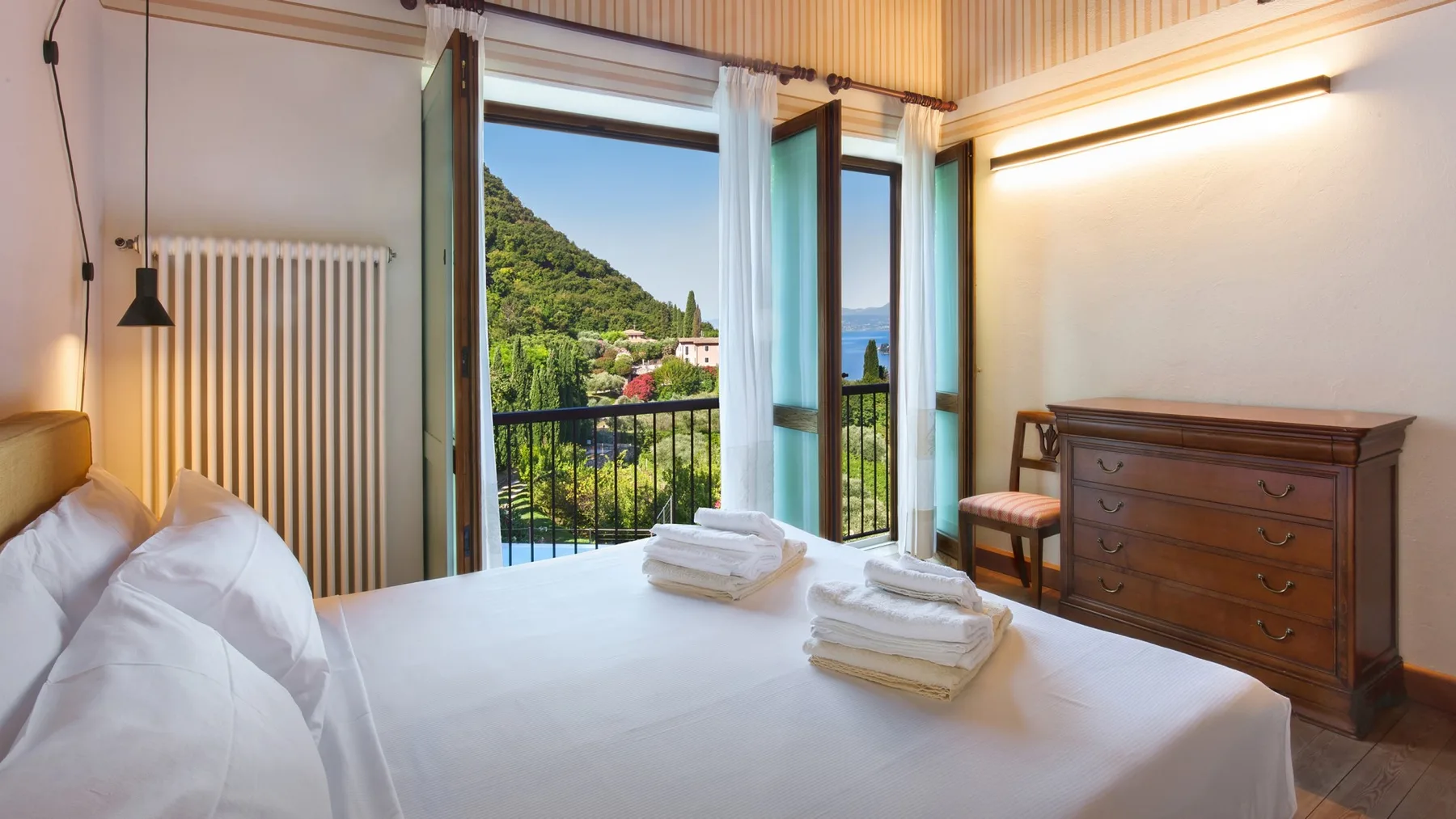 A bright bedroom with a bed facing open balcony doors and a green hillside view.