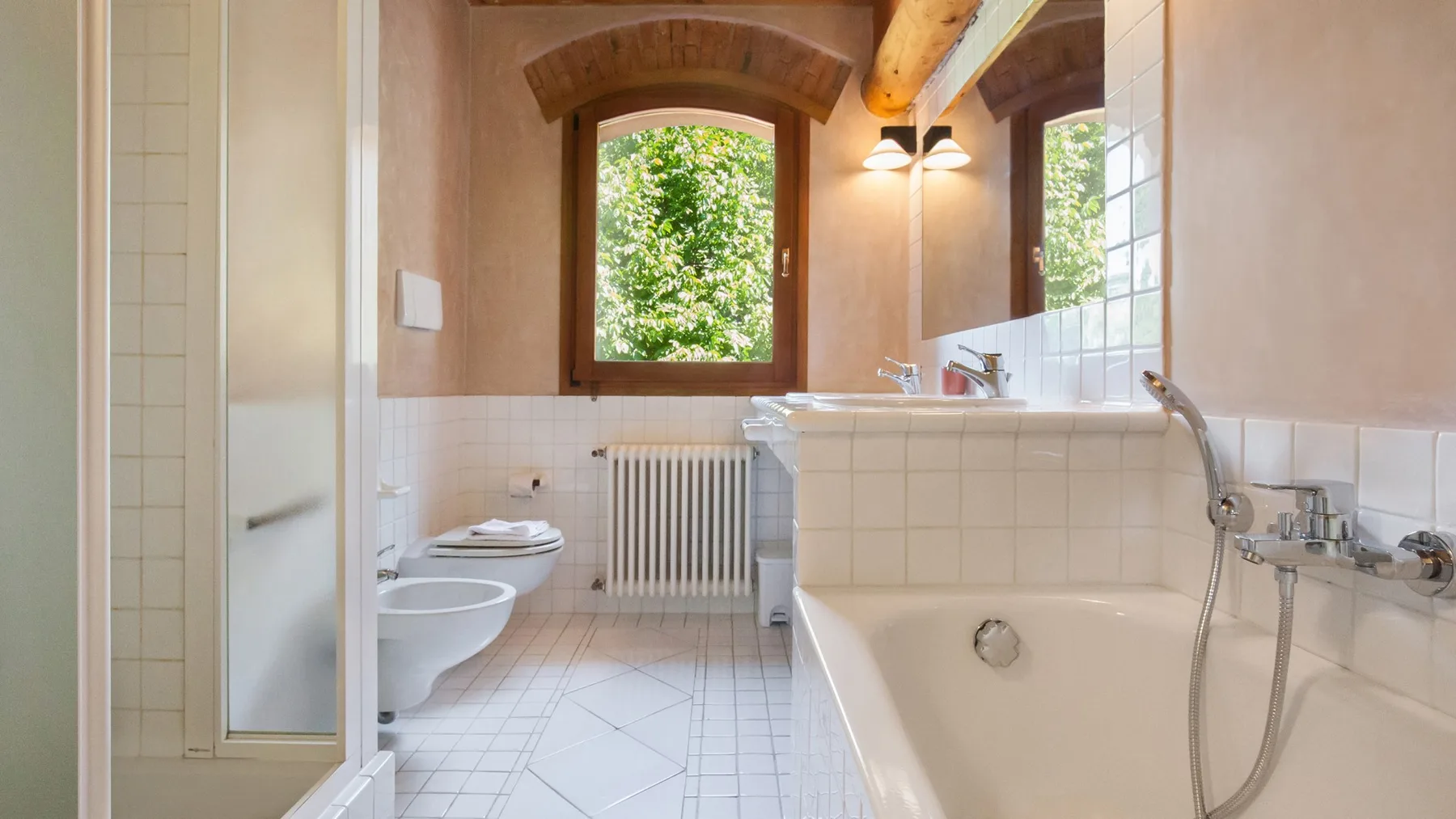 A bright bathroom with a bathtub, shower fixtures, and a window overlooking greenery.