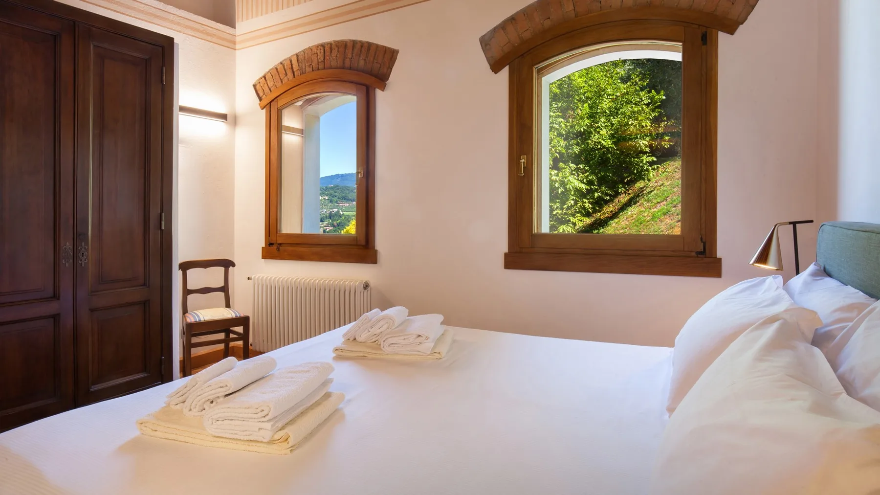 A bedroom with a bed, folded towels, and windows overlooking greenery.