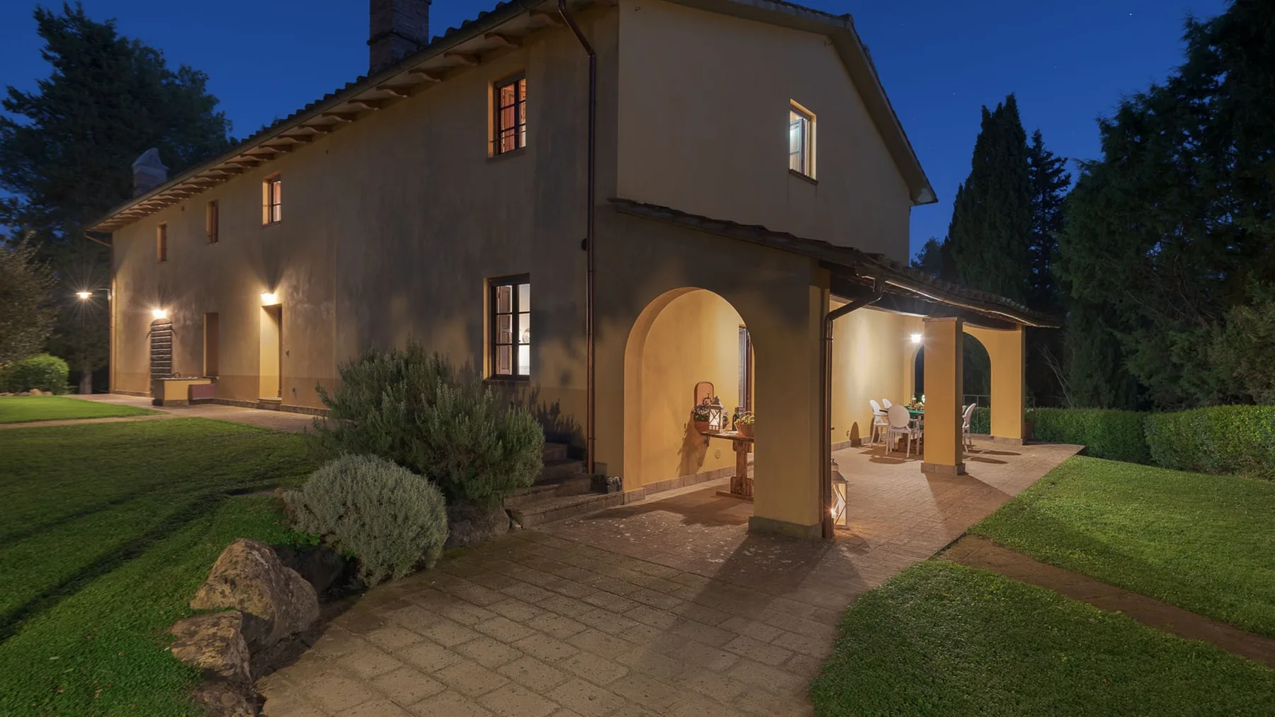 Una casa iluminada se alza junto a un patio pavimentado y un jardín al atardecer.
