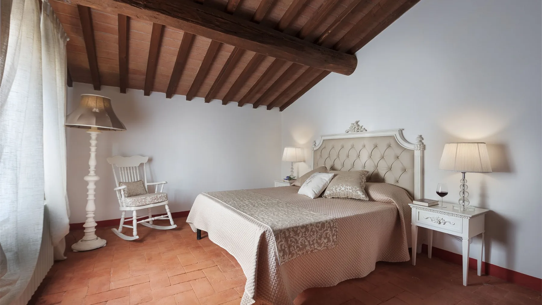 Un dormitorio con una cama doble, mesitas de noche y una mecedora bajo vigas de madera vistas.