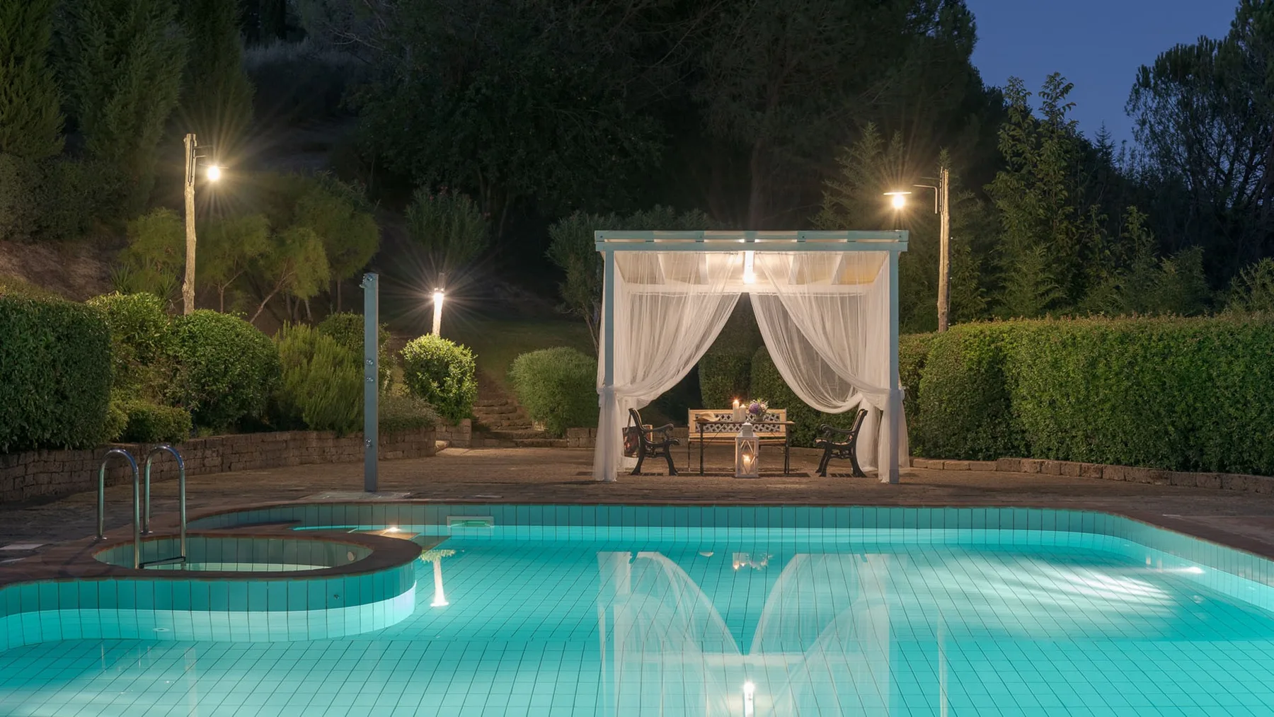 Una piscina exterior iluminada está frente a un pabellón de jardín con cortinas.