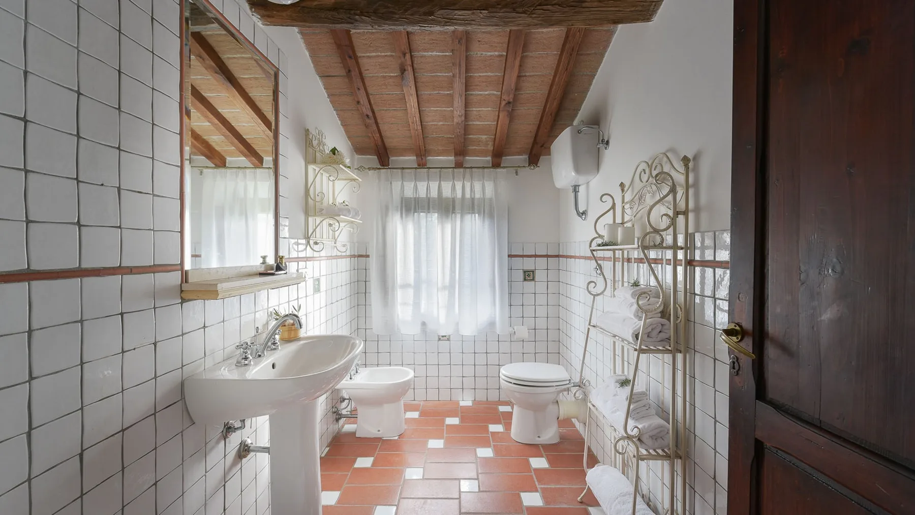 Un baño alicatado con lavabo, inodoros y una ventana con cortina.
