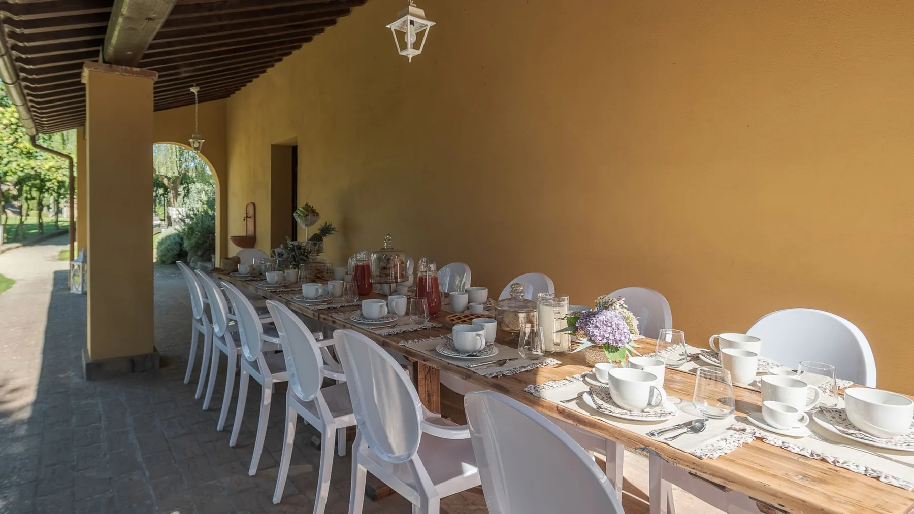 Una larga mesa está puesta con vajilla bajo una terraza exterior techada.