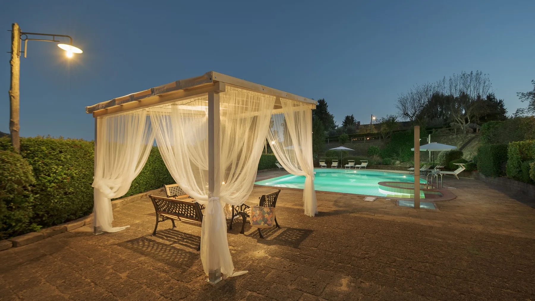 Una cabaña junto a la piscina, con cortinas translúcidas, está junto a una piscina iluminada.
